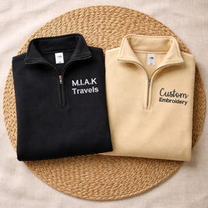 May include: A black quarter-zip sweatshirt with white text reading M.I.A.K Travels and a beige quarter-zip sweatshirt with black text reading Custom Embroidery. Both garments feature a high collar, a metal zip, and a Fruit of the Loom label.