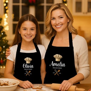 May include: Two people wearing black aprons with a chef's hat design and personalized names. The aprons have the text "Head Chef" and crossed utensils. A Christmas tree with lights and ornaments is in the background.