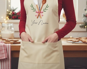 Può includere: Grembiule color crema con un'ampia tasca frontale. Il grembiule presenta un disegno ad acquerello di una spatola, una frusta e un cucchiaio di legno, legati con un nastro rosa e circondati da foglie verdi. Le parole "Your Name" sono scritte in corsivo.