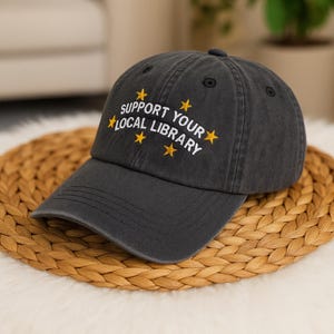 May include: A dark grey baseball cap with the text "SUPPORT YOUR LOCAL LIBRARY" embroidered in white and yellow. The hat has a curved peak and is on a woven mat.