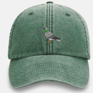 May include: A green baseball cap with a worn appearance. The cap displays an embroidered image of a pigeon in grey, white, and green. The cap has a curved peak and an adjustable strap.