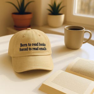 Peut inclure: Casquette beige avec le texte brodé "Born to read books forced to read emails". La casquette est posée sur une table blanche à côté d'un livre ouvert et d'une tasse beige. Deux plantes en pot sont en arrière-plan.