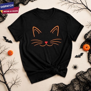 May include: A black short-sleeved t-shirt featuring a minimalist orange line art graphic of a cat face. The design includes two pointed ears, three whiskers on each side, and a small red heart shape positioned in the centre as the cat's nose.