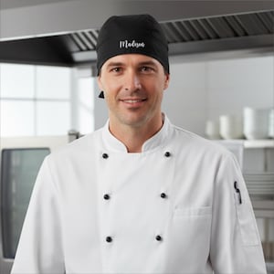 以下が含まれることがあります： 白い筆記体で「Madison」という名前が入った黒いシェフの頭巾。 黒いボタンが付いた白いシェフコートを着た人がかぶっています。