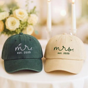 Puede incluir: Dos gorras de béisbol, una verde oscuro y otra beige, una al lado de la otra. La gorra verde oscuro tiene la palabra "mr." y el texto "est. 2025" en escritura blanca. La gorra beige tiene la palabra "mrs." y el texto "est. 2025" en escritura verde.