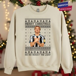 Puede incluir: Jersey navideño beige con un diseño festivo. El jersey presenta el texto "MERRY CHRISTMAS" y "HO HO HOWAY THE LADS!". Incluye un gráfico de una persona con una camiseta a rayas blancas y negras. El jersey tiene cuello redondo.