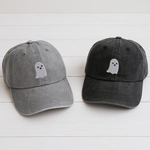 以下が含まれることがあります： グレーと黒の2つの野球帽が表示されています。各帽子には、前面に白い刺繍のゴーストデザインがあります。帽子は、カジュアルウェアに最適な、クラシックで調整可能なフィット感です。柔らかく耐久性のある素材で作られています。