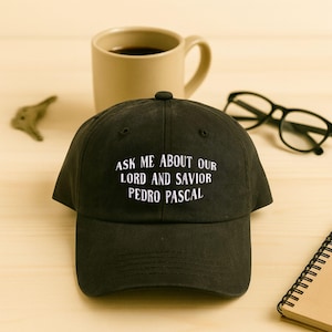May include: Black baseball cap with the embroidered text "ASK ME ABOUT OUR LORD AND SAVIOR PEDRO PASCAL" in white. A coffee mug, glasses, and a notepad are in the background.