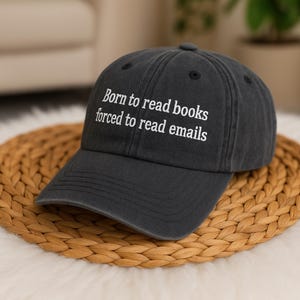 Może przedstawiać: Ciemnoszara czapka bejsbolowa z białym napisem "Born to read books forced to read emails". Czapka leży na okrągłej, plecionej podkładce.