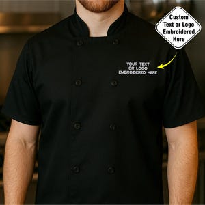 Op de afbeelding: Zwarte koksjas met korte mouwen en een opstaande kraag. De jas heeft zes zwarte knopen en de tekst "YOUR TEXT OR LOGO EMBROIDERED HERE" geborduurd op de borst. Een ruitvormige afbeelding leest "Custom Text or Logo Embroidered Here".