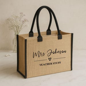 May include: Tan tote bag with black trim and handles. The bag features the text "Mrs Johnson" above a heart and the words "TEACHER STUFF" in black script. The bag is made of a woven material.