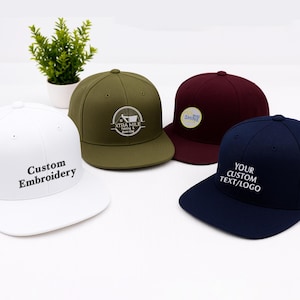 May include: Four baseball caps in white, olive green, burgundy, and navy blue. The white cap has "Custom Embroidery" in black. The olive green cap has a white logo. The burgundy cap has a yellow and blue logo. The navy cap has "YOUR CUSTOM TEXT/LOGO" in white.