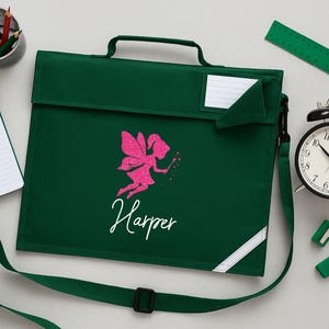 May include: A dark green school satchel with a pink glitter fairy design and the name "Harper" in white script. Includes a shoulder strap, handle, and a white label area. A green ruler, pencils, and a clock are also visible.