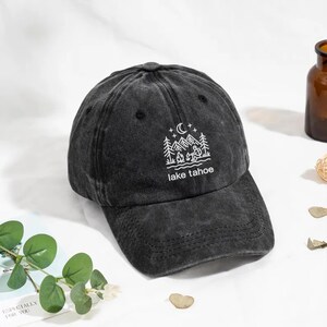 May include: A black, distressed baseball cap with a curved peak. The cap features a white graphic of a mountain scene with the text "lake tahoe". The cap is made of a soft, worn material.