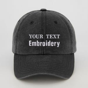 May include: A dark grey baseball cap featuring the white embroidered text "YOUR TEXT Embroidery". The cap has a curved peak and a button on top. The hat is made of a soft, worn material.