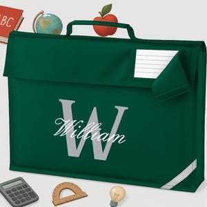 Puede incluir: Un bolso escolar verde oscuro con el nombre "William" y una gran "W" plateada. El bolso tiene un asa y una ranura para etiquetas con nombre. Ilustraciones escolares, incluyendo un libro rojo, un globo y una manzana, rodean el bolso.