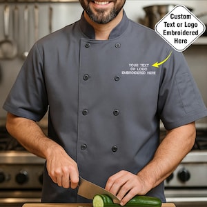 Op de afbeelding: Een antracietgrijze koksjas met korte mouwen en zwarte knopen. De jas heeft een klein zakje op de linkermouw en de tekst "YOUR TEXT OR LOGO EMBROIDERED HERE" geborduurd op de borst. De afbeelding bevat ook de tekst "Aangepaste tekst of logo hier geborduurd".