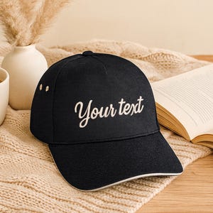 Könnte beinhalten: Eine schwarze Baseballkappe mit gebogenem Schirm und dem gestickten Text "Your text" in Schreibschrift. Die Kappe hat einen weißen Rand entlang des Schirms und ist vor einem neutralen Hintergrund platziert.