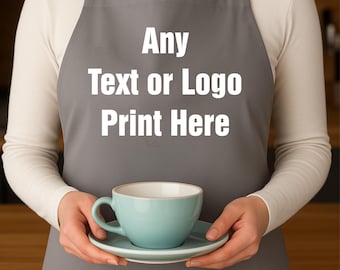 May include: A gray apron with the text "Any Text or Logo Print Here" in white. The apron has a pocket and is worn over a white long-sleeved shirt. A person holds a teal teacup and saucer.