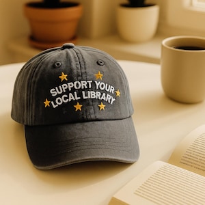 Peut inclure: Casquette de baseball gris foncé avec le texte brodé "SUPPORT YOUR LOCAL LIBRARY" en blanc et des étoiles jaunes. La casquette est posée sur une surface claire, avec une tasse à café et un livre ouvert à proximité.