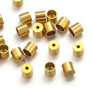 Puede incluir: Un conjunto de pequeñas cuentas cilíndricas de latón con un agujero en el centro. Las cuentas están dispersas sobre una superficie blanca, y la iluminación resalta su brillo metálico. Estos componentes se utilizan probablemente en la fabricación de joyas u otras manualidades.