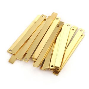Puede incluir: Un conjunto de barras metálicas doradas y brillantes, con bordes redondeados y un pequeño agujero en un extremo. Las barras son rectangulares y parecen estar hechas de un material reflectante. Están dispuestas de forma ligeramente superpuesta.