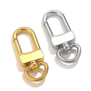 May include: Two metal swivel clasps, one gold-toned and one silver-toned, are displayed against a white background. Each clasp has a rectangular base, a curved top, and a small ring at the bottom. The gold clasp is on the left, and the silver clasp is on the right.