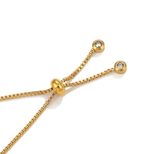 Könnte beinhalten: Nahaufnahme eines goldfarbenen Armbands mit einem verschiebbaren Perlen und zwei Endstücken. Jedes Endstück hat einen kleinen, klaren, runden Stein. Die Kette des Armbands hat ein Box-Link-Design. Der Hintergrund ist weiß.