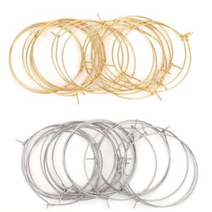 Puede incluir: Dos juegos de componentes de pendientes de aro metálicos. Uno es dorado y el otro plateado. Cada juego contiene múltiples aros circulares con pequeñas extensiones de alambre para sujetar ganchos u otros adornos. Los aros están dispuestos de forma ordenada.