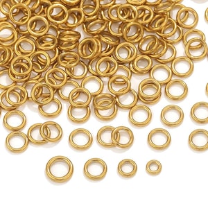 May include: A collection of small, gold-coloured metal rings. The rings are round with a consistent thickness and a smooth, shiny surface. They are scattered across a white background, with varying sizes.