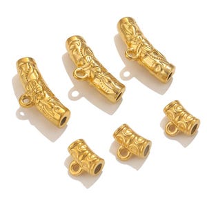 May include: Six gold-coloured metal tube beads with floral and swirl patterns. Each bead has a small loop on one end and a hole on the other. The beads are arranged on a white surface.