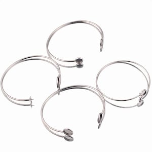 May include: Four silver-colored, open-ended bracelets. Each bracelet has a double-band design and small, oval-shaped end pieces. The bracelets are displayed on a reflective surface, creating a mirrored effect. The overall design is minimalist and modern.