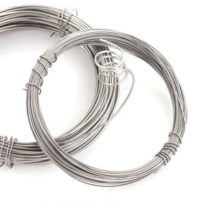 Può includere: Due bobine di filo color argento sono mostrate su sfondo bianco. Il filo è avvolto strettamente in forme circolari, con una bobina parzialmente avvolta e fissata. Il filo sembra sottile e flessibile, adatto per l'artigianato o la creazione di gioielli.