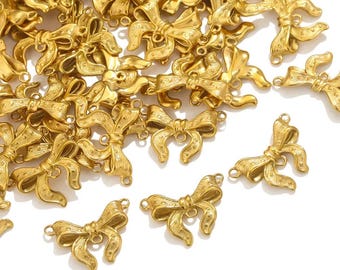 May include: A collection of gold-colored bow charms. Each charm features a detailed bow design with textured surfaces and small loops for attaching to jewelry or other crafts. The charms are scattered on a white surface.
