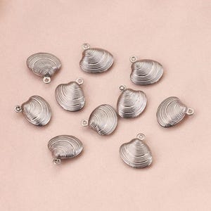Peut inclure: Un ensemble de breloques argentées en forme de coquillage. Chaque breloque présente un motif de coquillage détaillé et une petite boucle pour l'attacher à des bijoux ou à d'autres créations. Les breloques sont disposées sur une surface rose clair.