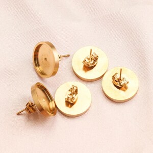 May include: Gold-colored earring blanks with posts and backs. The earring blanks are round with a flat surface for attaching designs. The posts are straight and the backs are butterfly style. The earrings are displayed on a light pink fabric.