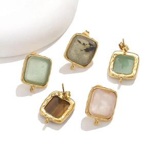 Op de afbeelding: Vijf goudkleurige oorbellen met rechthoekige edelstenen in verschillende kleuren: lichtgroen, grijs, bruin, roze en donkergroen. Elke oorbel heeft een unieke steen en een gouden frame.