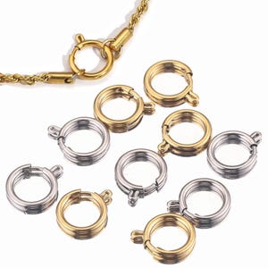 Könnte beinhalten: Eine Sammlung von gold- und silberfarbenen Federringverschlüssen, mit einer goldenen Kette im Hintergrund. Die Verschlüsse sind rund mit einem kleinen Hebel zum Öffnen und Schließen. Die Verschlüsse werden für die Schmuckherstellung verwendet.