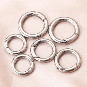 Peut inclure: Six fermoirs ronds argentés brillants avec une ouverture à charnière. Les fermoirs sont disposés sur un tissu rose clair. Les fermoirs sont en métal et ont une surface lisse et réfléchissante. Ils sont conçus pour la fabrication de bijoux ou d'autres projets d'artisanat.
