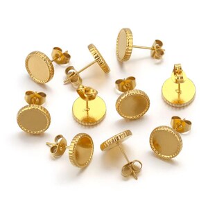 May include: A collection of gold-colored, round stud earring components. Each earring has a flat, circular base with a textured edge and a post for insertion. The earrings are scattered on a white surface.