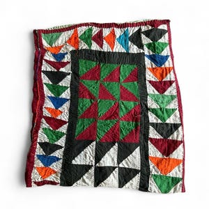 May include: A colorful, handmade quilt with a patchwork design. The quilt features a central square of red and green triangles, framed by a black border. The edges are adorned with rows of triangles in various colors, including orange, blue, and green. The quilt has a red border.
