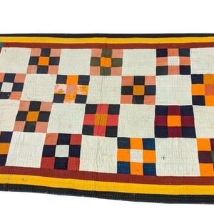 May include: A patchwork quilt with a repeating pattern of squares in white, orange, red, and navy blue. The quilt has a yellow and black border. The quilt is a handmade textile.