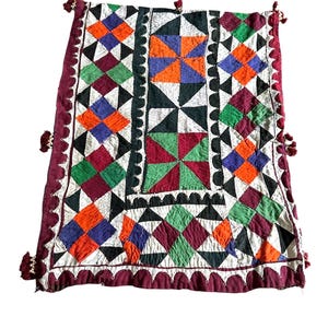 May include: A colorful, geometric patterned textile with a burgundy border and decorative tassels. The design features squares and triangles in shades of orange, blue, green, and burgundy, set against a white background. The edges are scalloped.