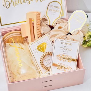 May include: A pink gift box with gold detailing, inscribed with "Will You Be My Bridesmaid?", holds various items. These include hair accessories, cards, and a small container. The box contains a hair claw clip, scrunchies, and hair clips.