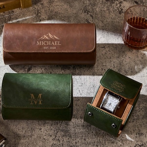 May include: Leather watch cases in brown and green. The brown case is embossed with "MICHAEL EST. 2026" and a mountain graphic. The green case displays the letter "M". A watch is displayed in a green case.