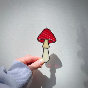 Puede incluir: Un adorno de hongo de vidriera con un sombrero rojo con puntos blancos y un tallo color crema. El hongo está delineado en negro y tiene un pequeño lazo para colgarlo. El hongo se sostiene frente a un fondo blanco.