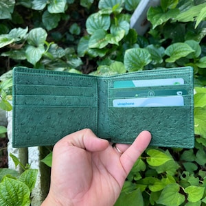 Puede incluir: Cartera de cuero verde abierta, con múltiples ranuras para tarjetas y una superficie texturizada. La cartera se sostiene abierta, revelando varias tarjetas en su interior. El color de la cartera es un verde intenso y rico.