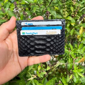 Puede incluir: Tarjetero negro con un patrón de piel de serpiente texturizada. La cartera es rectangular con múltiples ranuras para tarjetas, parcialmente llenas de tarjetas. El tarjetero se sostiene en una mano sobre un fondo de follaje verde.