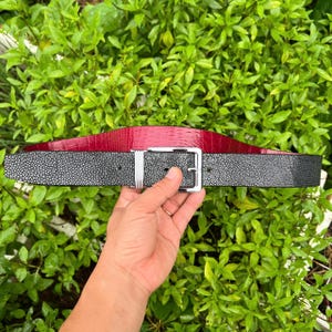 Puede incluir: Un cinturón reversible con un lado negro texturizado y un lado rojo con relieve de cocodrilo. El cinturón tiene una hebilla rectangular de color plateado. El cinturón se sostiene sobre un fondo de follaje verde.