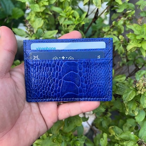 Puede incluir: Un tarjetero azul vibrante con un patrón texturizado y en relieve. La cartera tiene dos ranuras para tarjetas, una de las cuales contiene una tarjeta con el texto "vinaphone". El tarjetero se sostiene en una mano sobre un fondo de follaje verde.